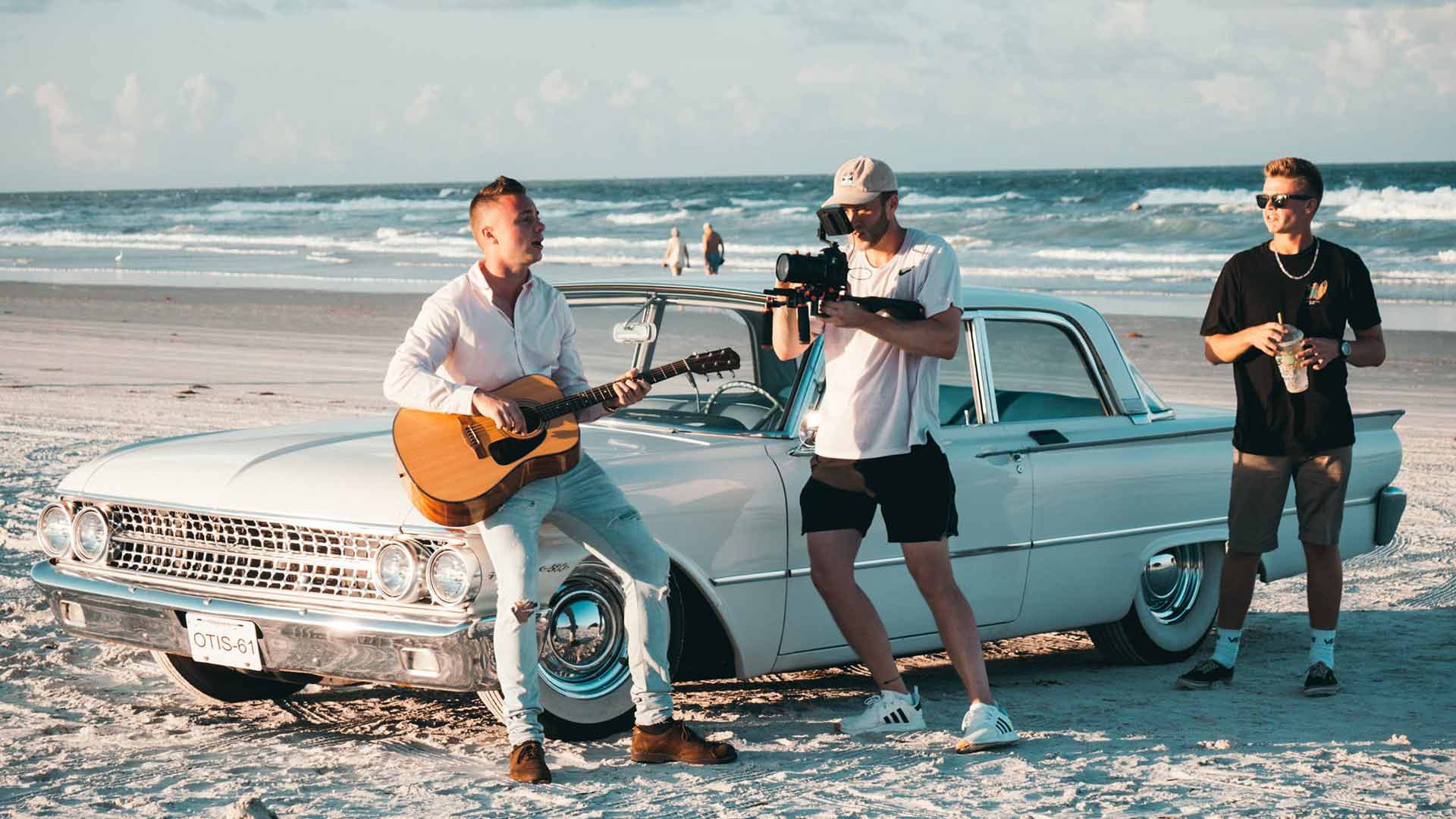 Musician playing guitar on a vintage Cadillac at the beach with filmmaker shooting footage for a music video -music video and short film ideas